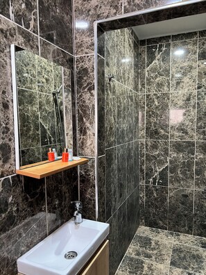 A bathroom with a marble-tiled shower, a white sink, and a wooden shelf with toiletries. - Mehmet Efendi Konagi Otel Restaurant (Akçaabat)