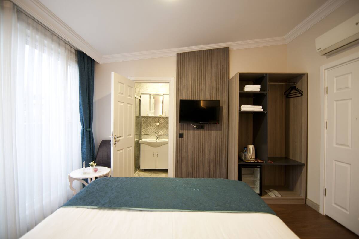 Une chambre d’hôtel comprenant un lit, une petite table avec une chaise, une salle de bain avec un lavabo et un miroir, un téléviseur fixé au mur et une armoire avec des vêtements suspendus.