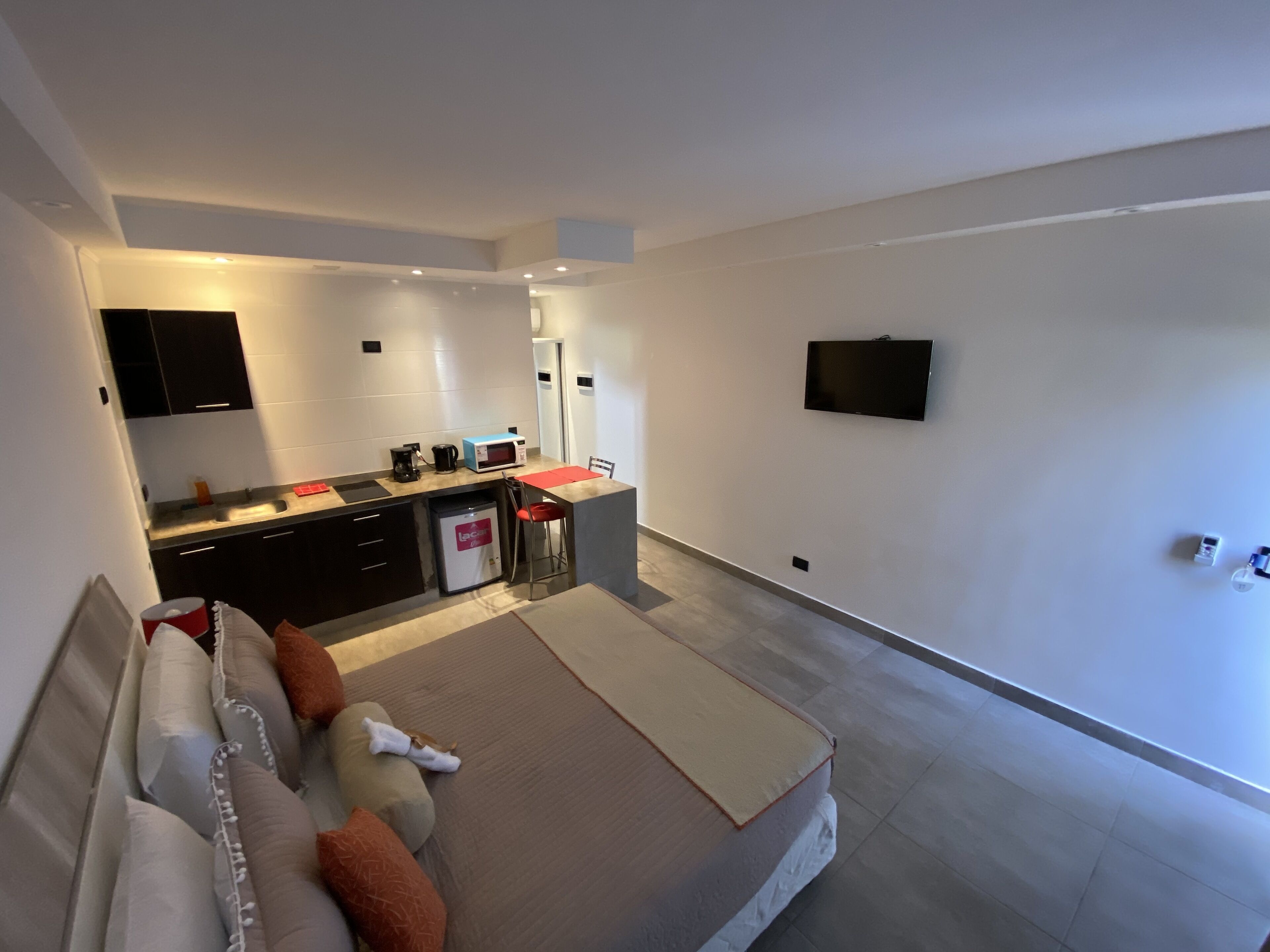 A modern hotel room with a kitchenette, a flat-screen TV, a sofa with cushions, and a large window.