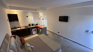 A modern hotel room with a kitchenette, a flat-screen TV, a sofa with cushions, and a large window.