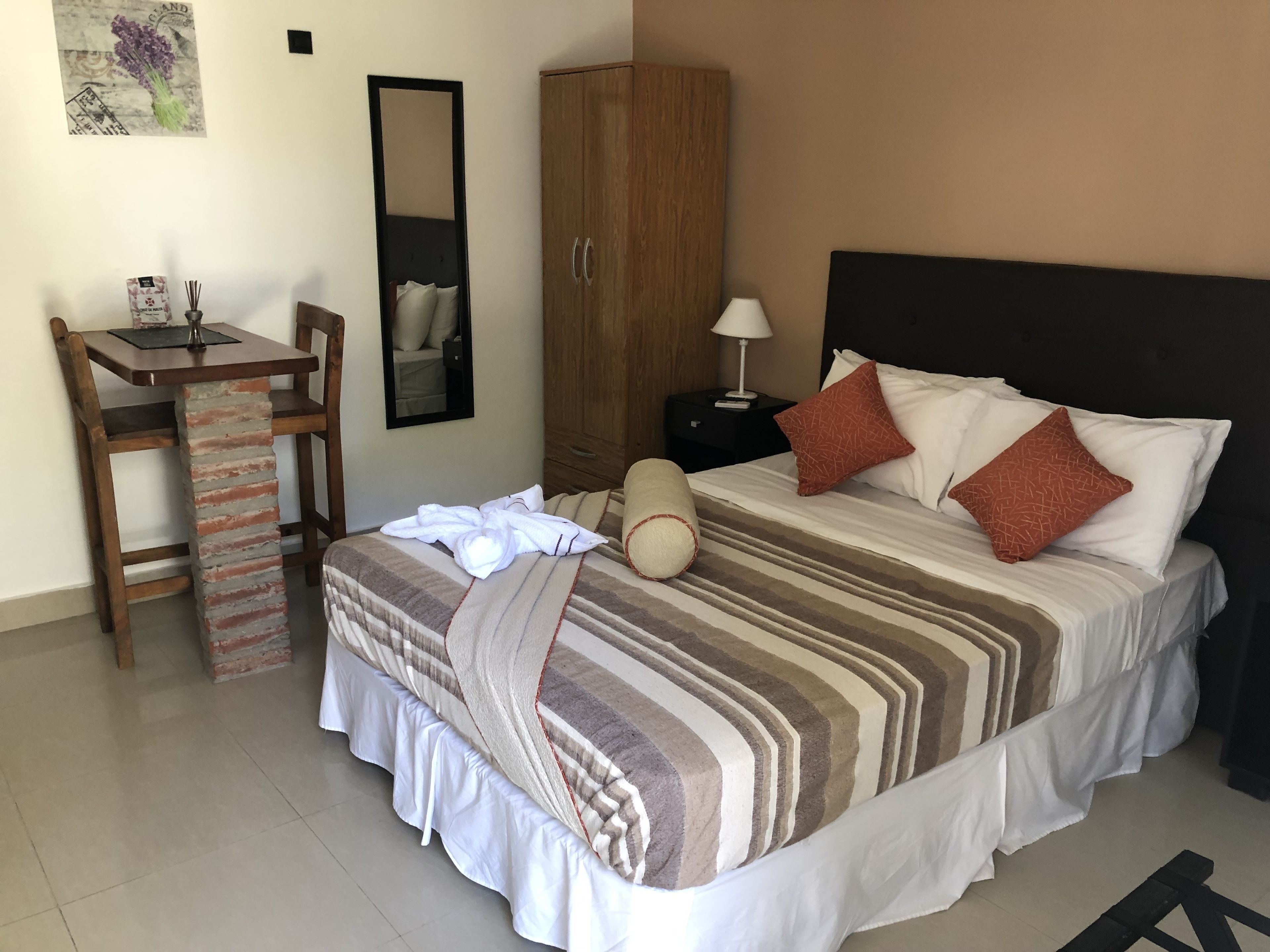A hotel room with a bed, a wooden desk, a wardrobe, a mirror, and a nightstand with a lamp.