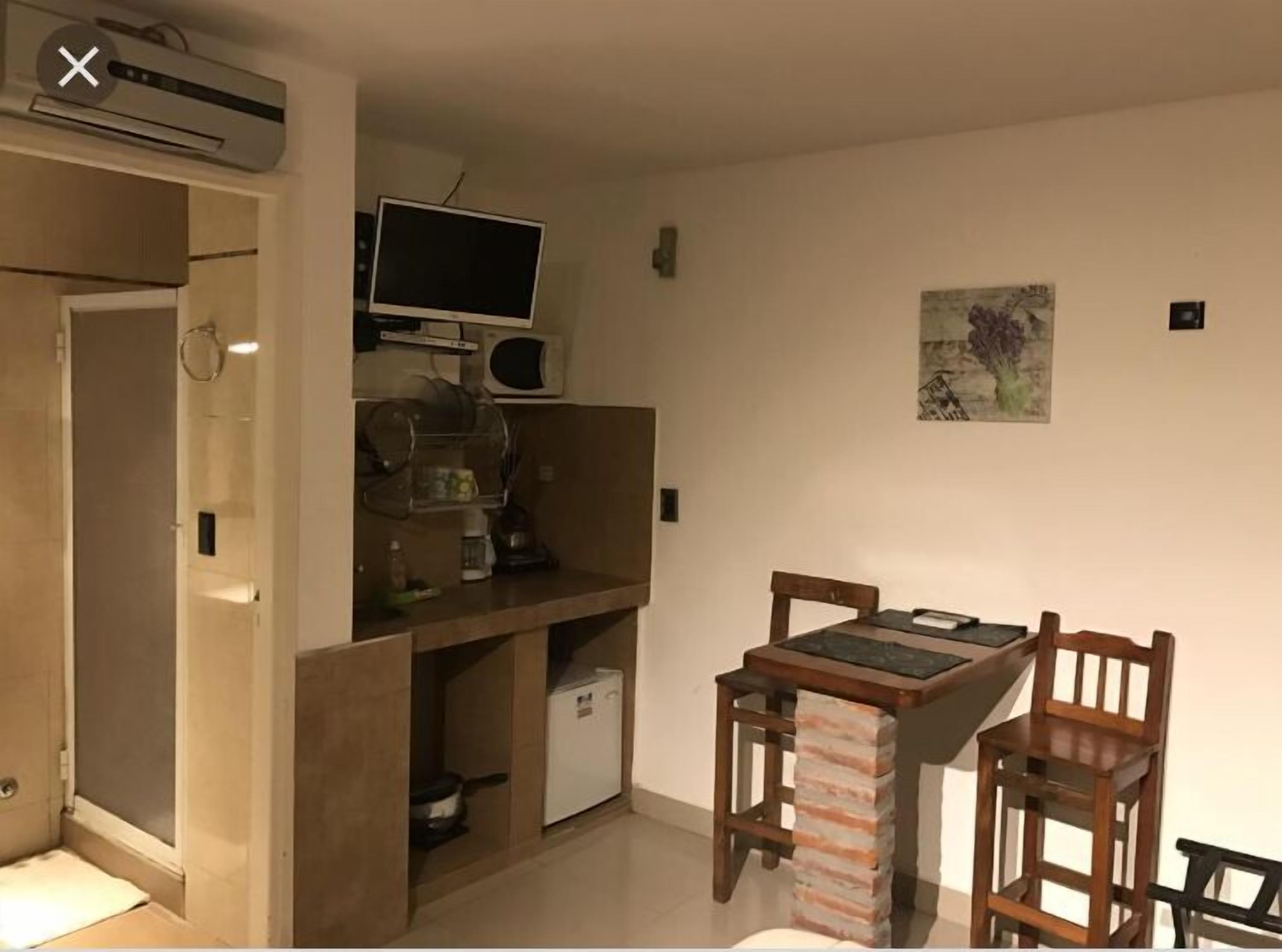 A compact kitchen with a built-in microwave, a small refrigerator, and a flat-screen TV. There is a wooden table and two chairs. A painting hangs on the wall.