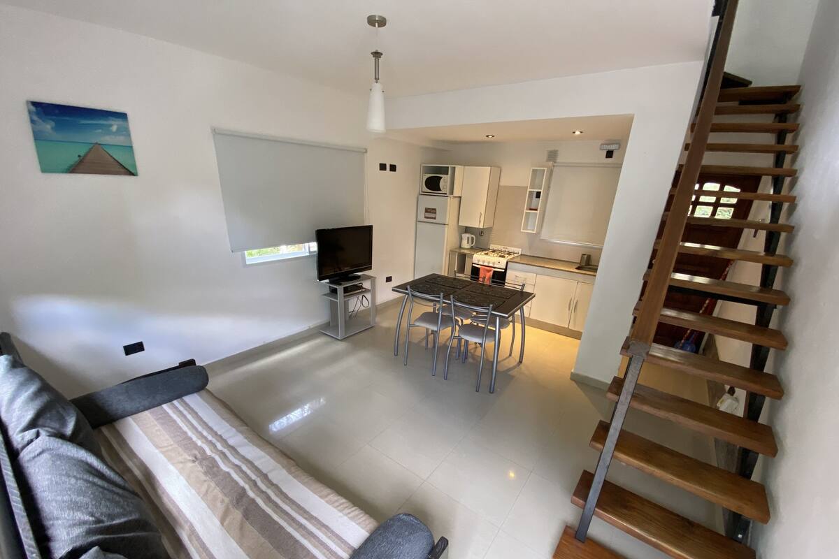 A modern living room with a sofa, a TV, and a dining area with a table and chairs. There is a kitchen visible in the background with a stove and cabinets.