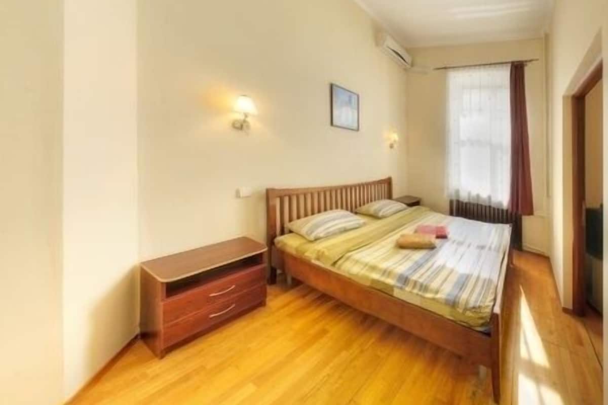 A bedroom with a wooden bed, a nightstand, a wall-mounted lamp, and a window with curtains.