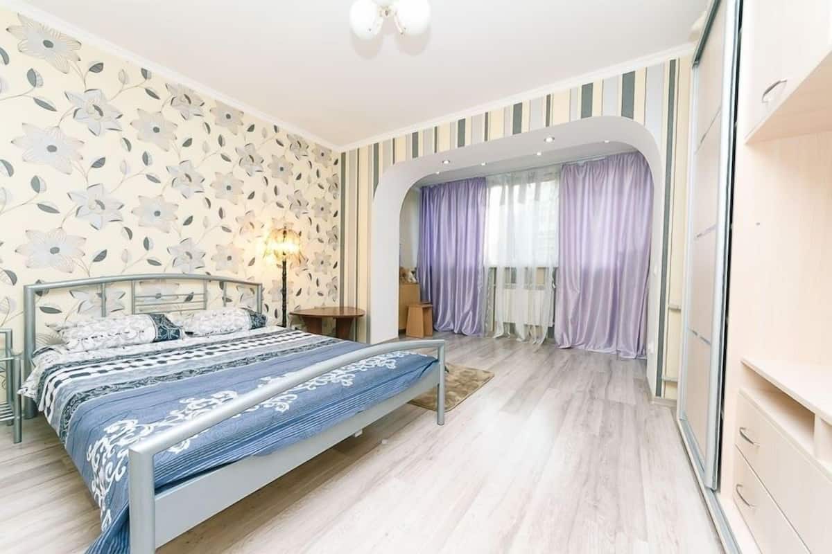 A bedroom with a bed, a nightstand, a lamp, a window with curtains, and a wardrobe.