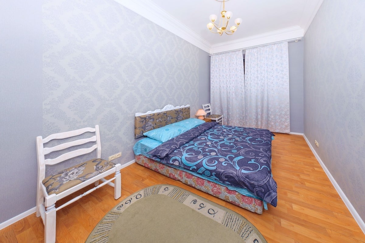 A bedroom with a bed, a chair, a nightstand, and a chandelier.