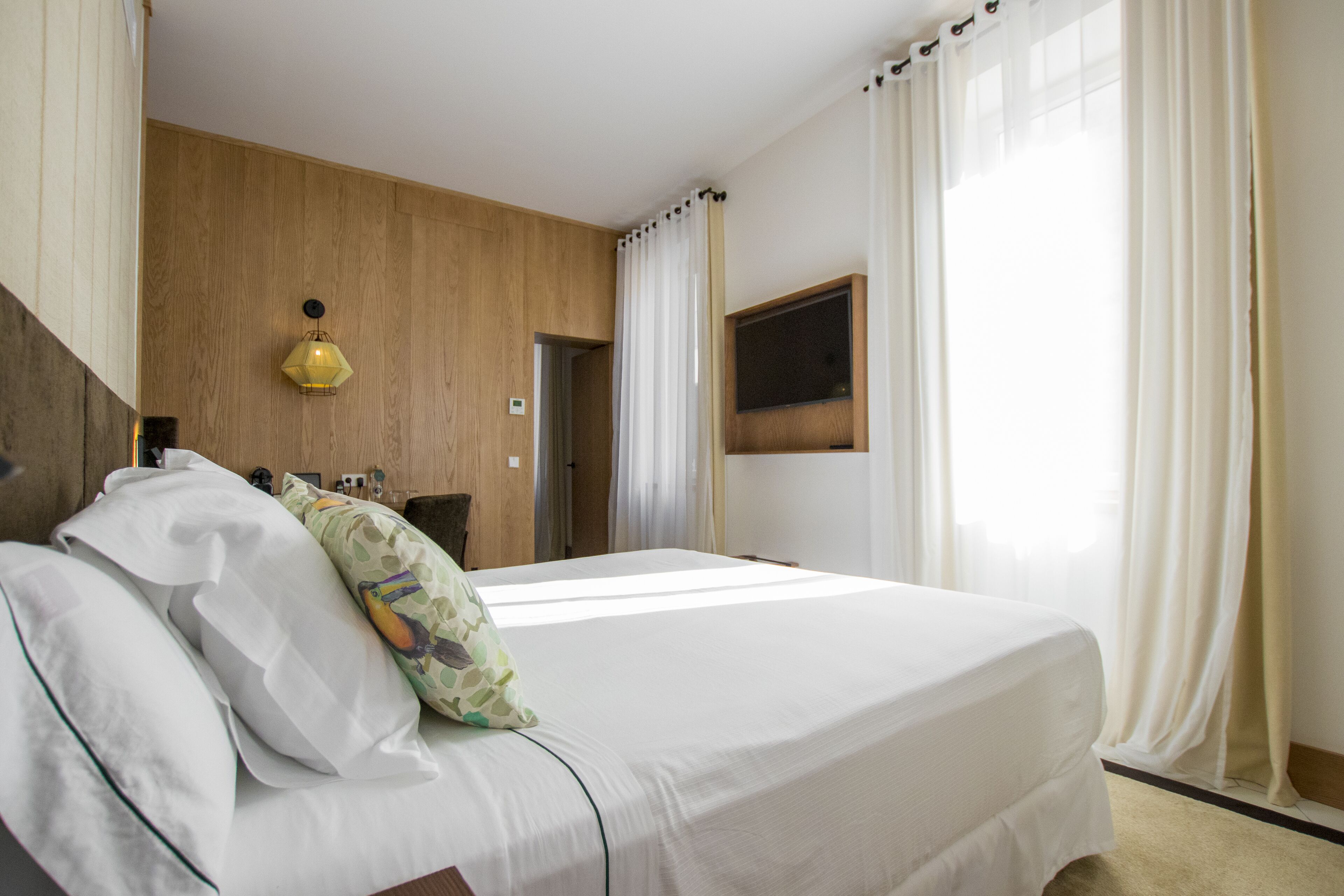 A hotel room with a bed, a wooden wall, a window with curtains, and a wall-mounted lamp.