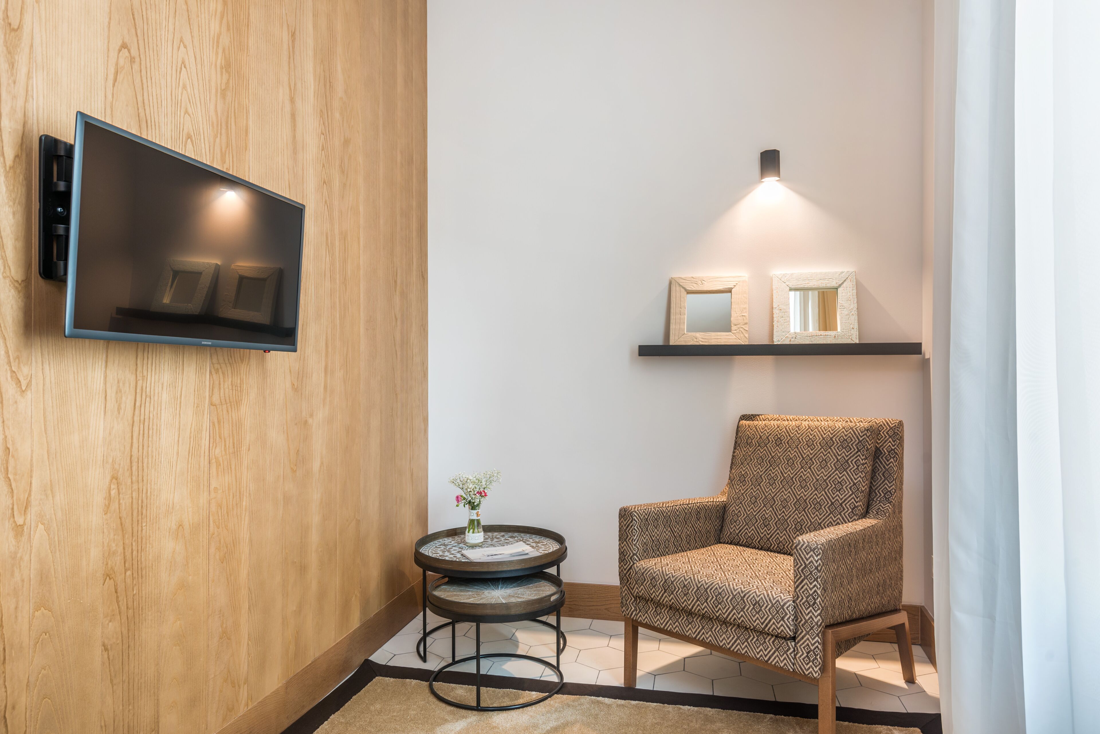 Ein modernes Zimmer mit montiertem Fernseher, einem gemusterten Sessel, einem kleinen runden Tisch mit einer Vase und einem Regal mit gerahmten Bildern.