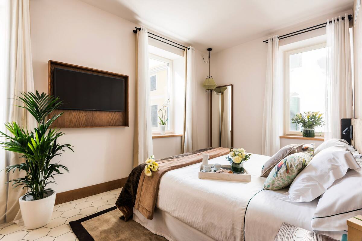 A hotel room with a bed, a TV mounted on the wall, a potted plant, a window with curtains, and a bedside table with a tray of items.