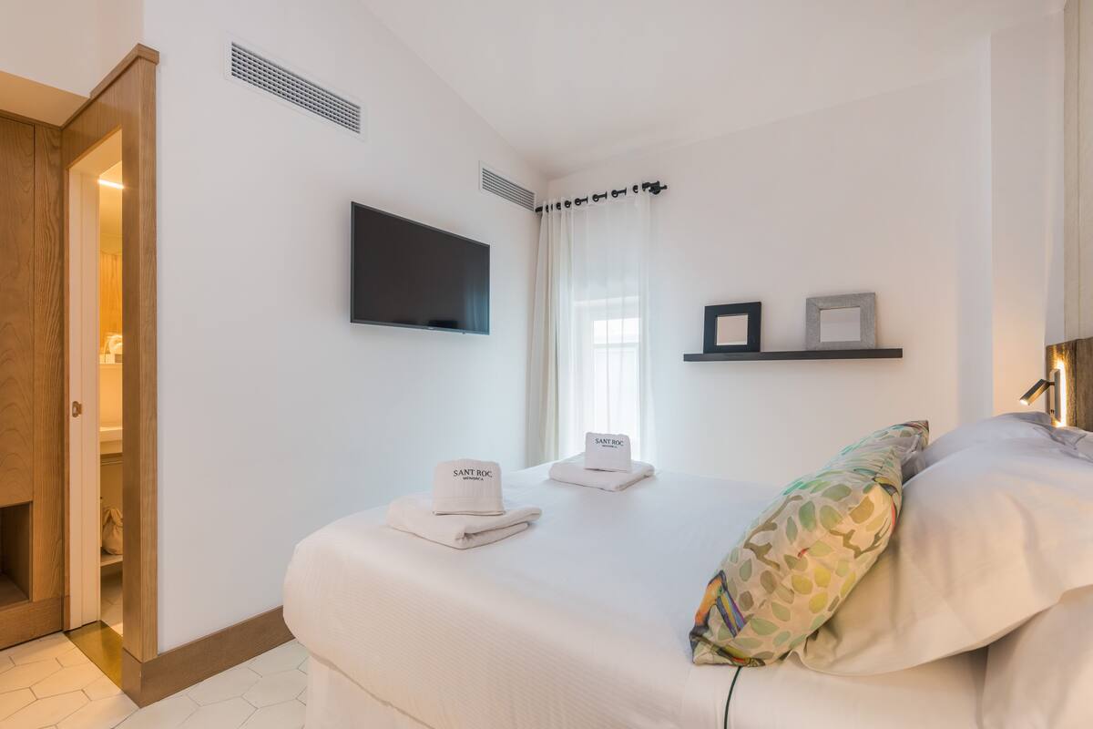 A hotel room with a bed, a TV mounted on the wall, a shelf with framed pictures, and a door leading to a bathroom.