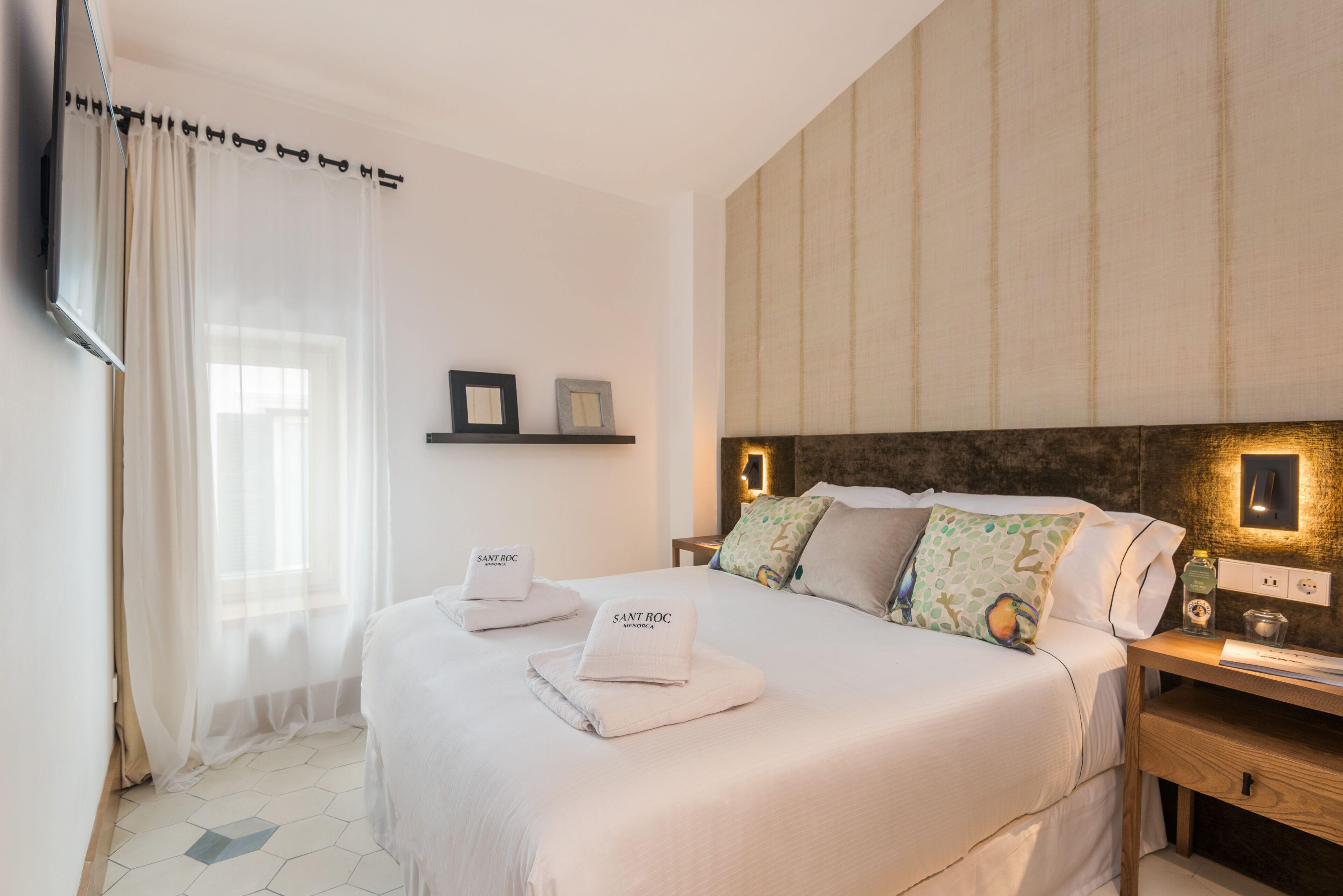 A hotel room with a large bed, a bedside table with a lamp, a wall-mounted TV, and a window with sheer curtains.