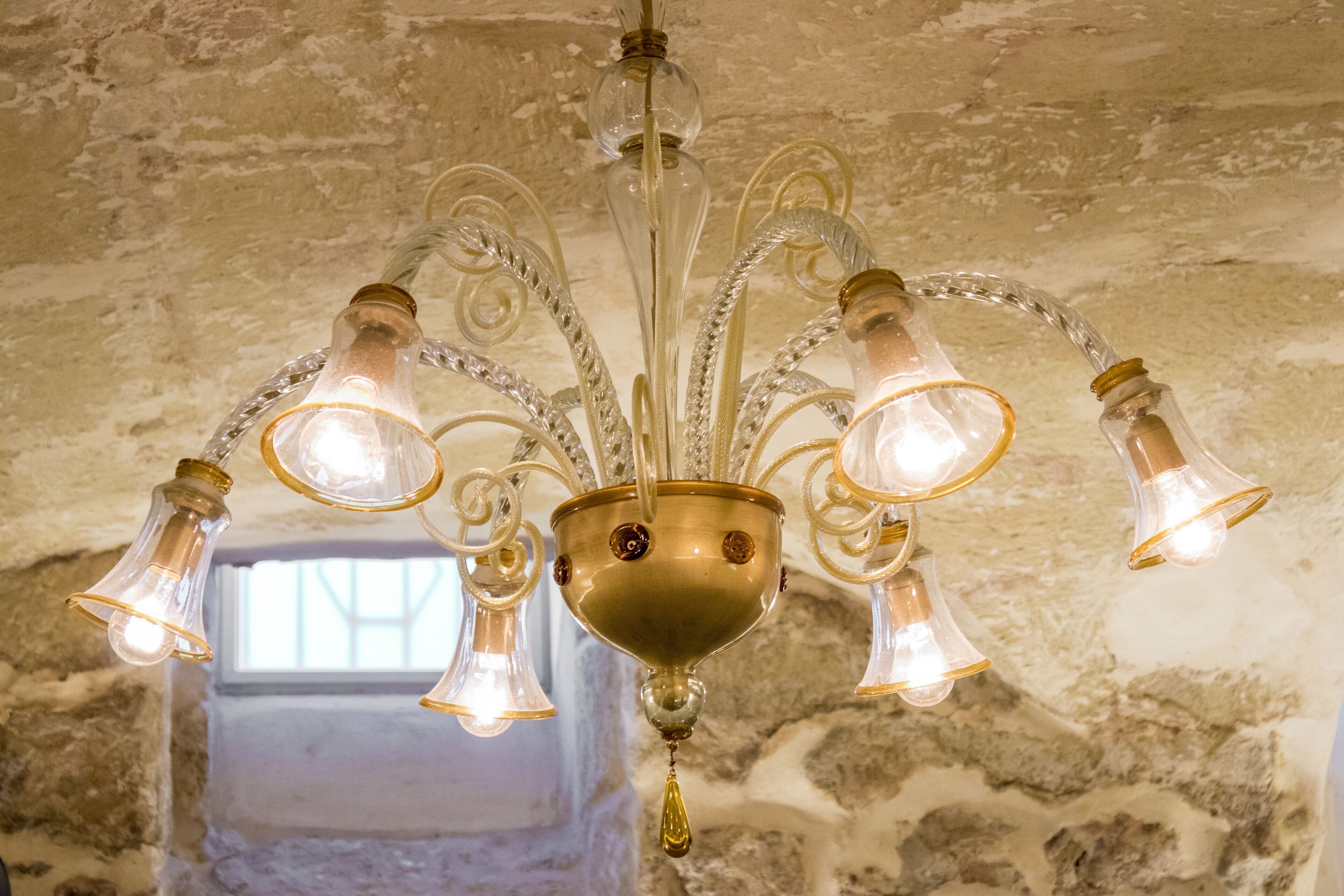 Een kroonluchter met meerdere glazen lampen en een messing voet, hangend in een kamer met een structuurwand van steen.