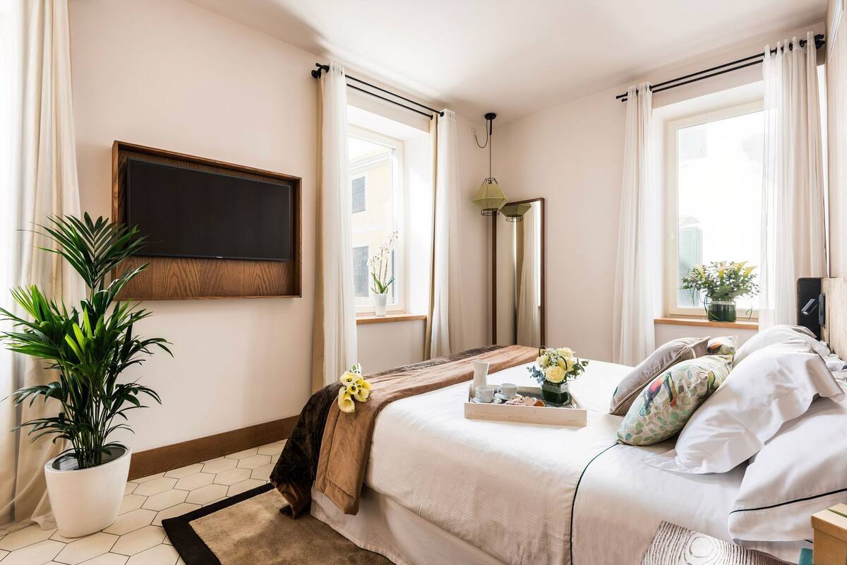 A hotel room with a bed, a TV mounted on the wall, a potted plant, and a window with curtains.