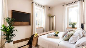 A hotel room with a bed, a TV mounted on the wall, a potted plant, and a window with curtains.