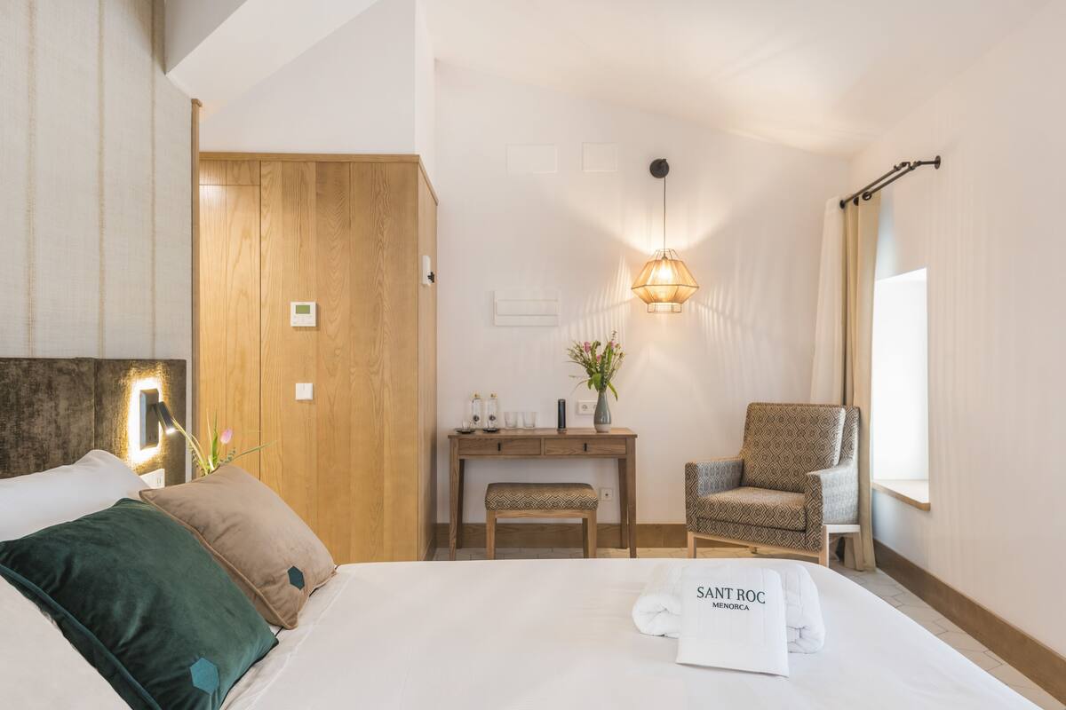 A hotel room with a bed, a wooden wardrobe, a desk with a chair, a bedside table with a vase, and a window with curtains.