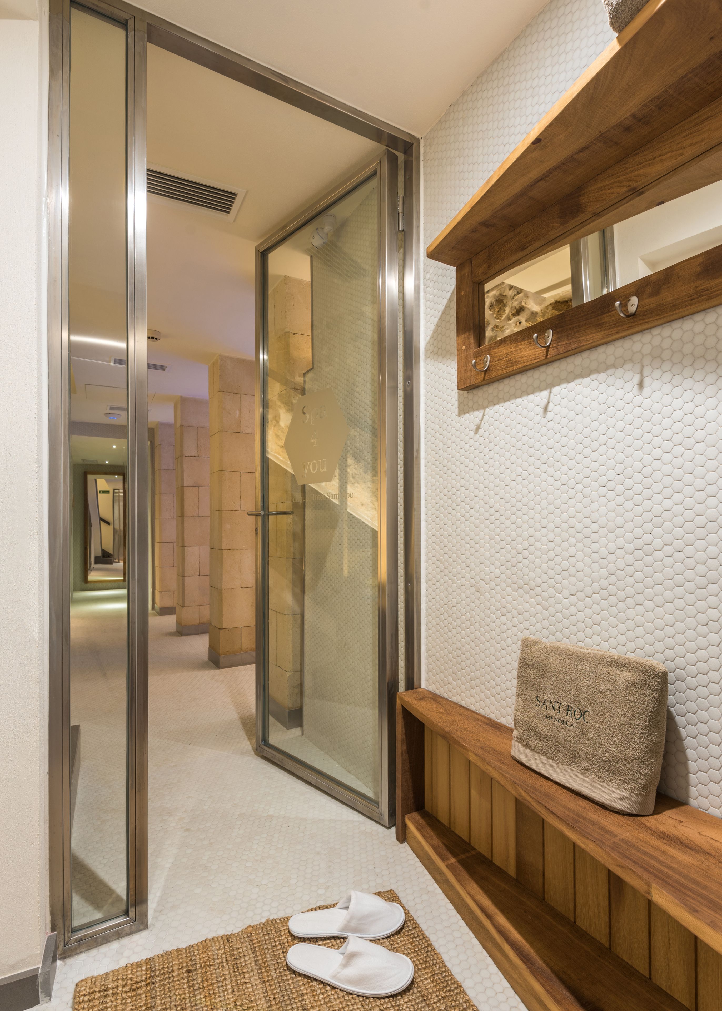 Un vestíbulo de hotel con un vidrio que da a un pasillo, un banco de madera con una toalla y una pared texturizada.