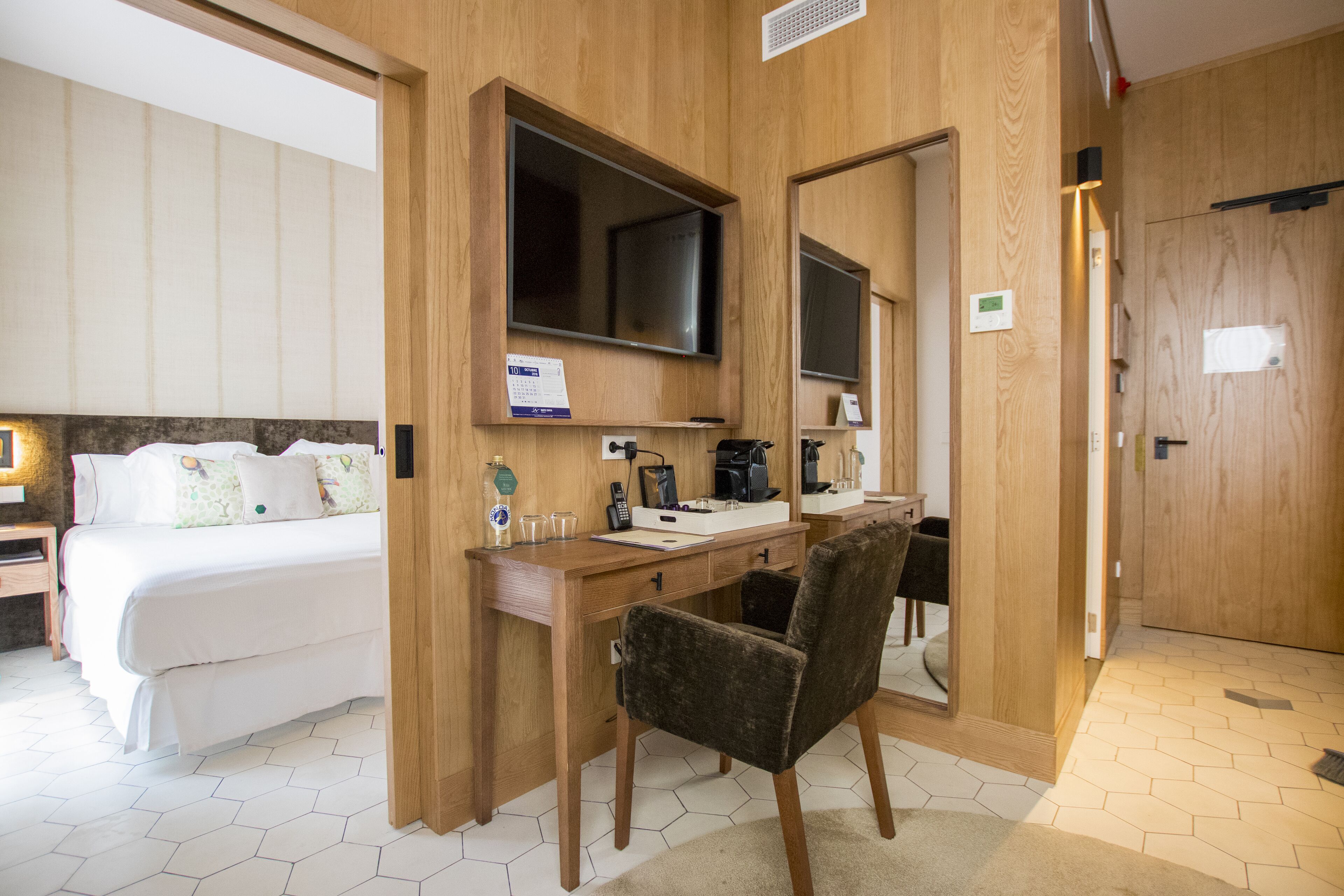 Habitación de hotel con cama, escritorio, silla, televisor y espejo.