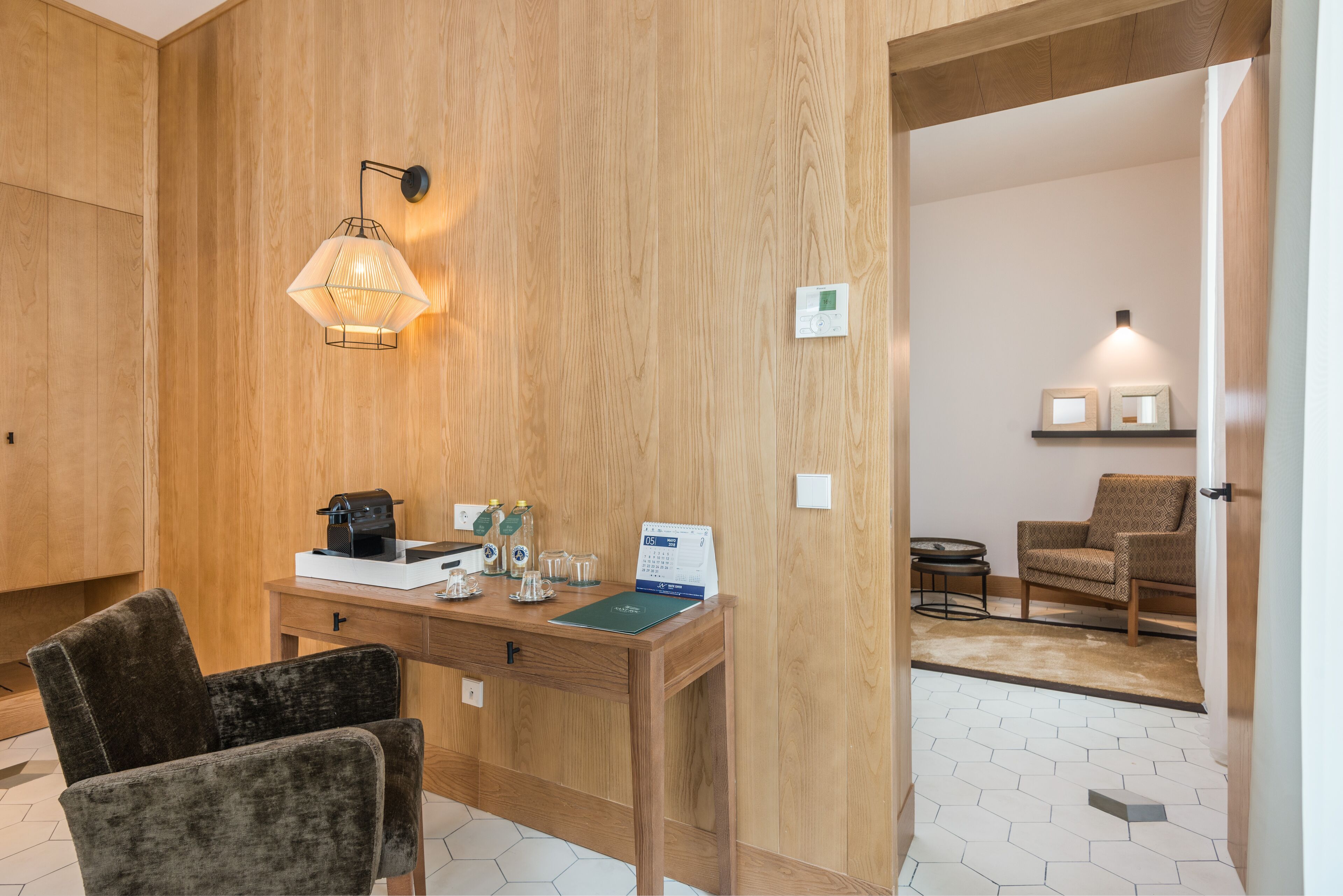 Habitación de hotel con pared de madera, un escritorio con cafetera, un calendario y dos sillas.