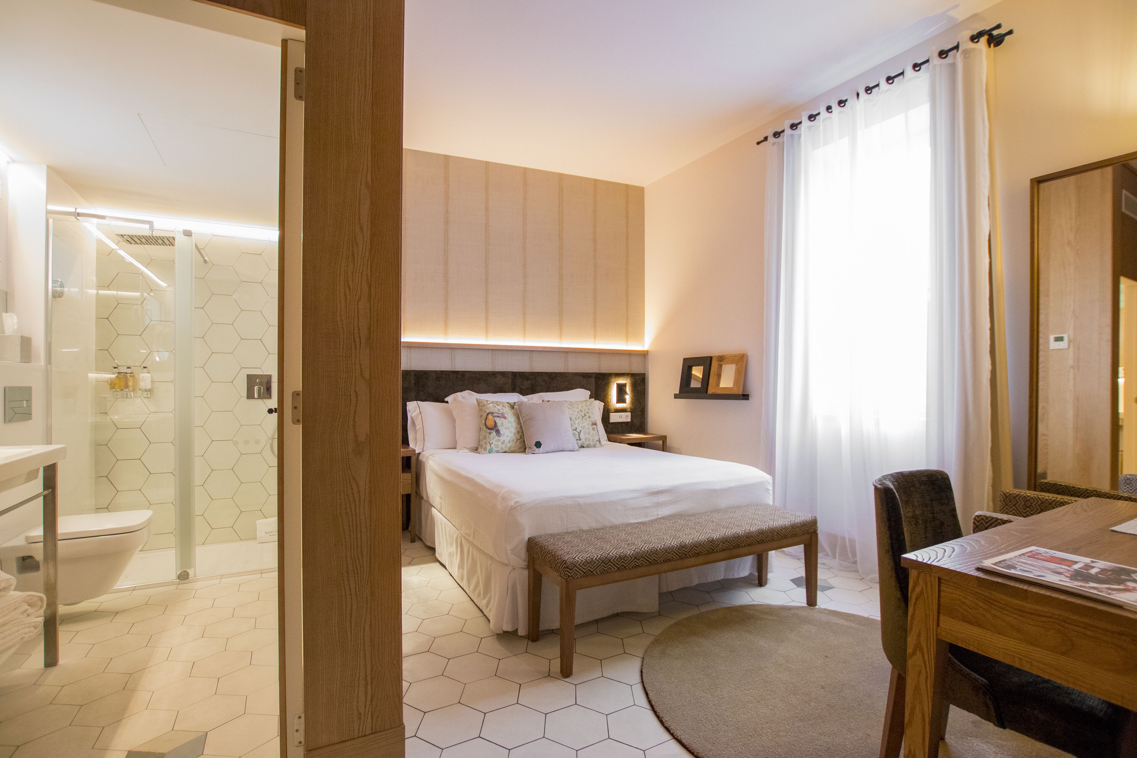 A hotel room with a bed, a bench, a table, and a bathroom visible through an open door.