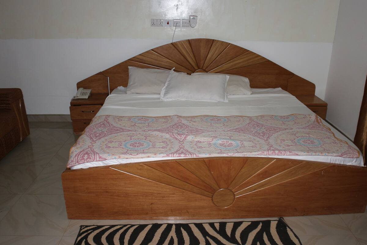 Uma cama de madeira com colcha estampada, uma mesa de cabeceira com um telefone e um criado-mudo.