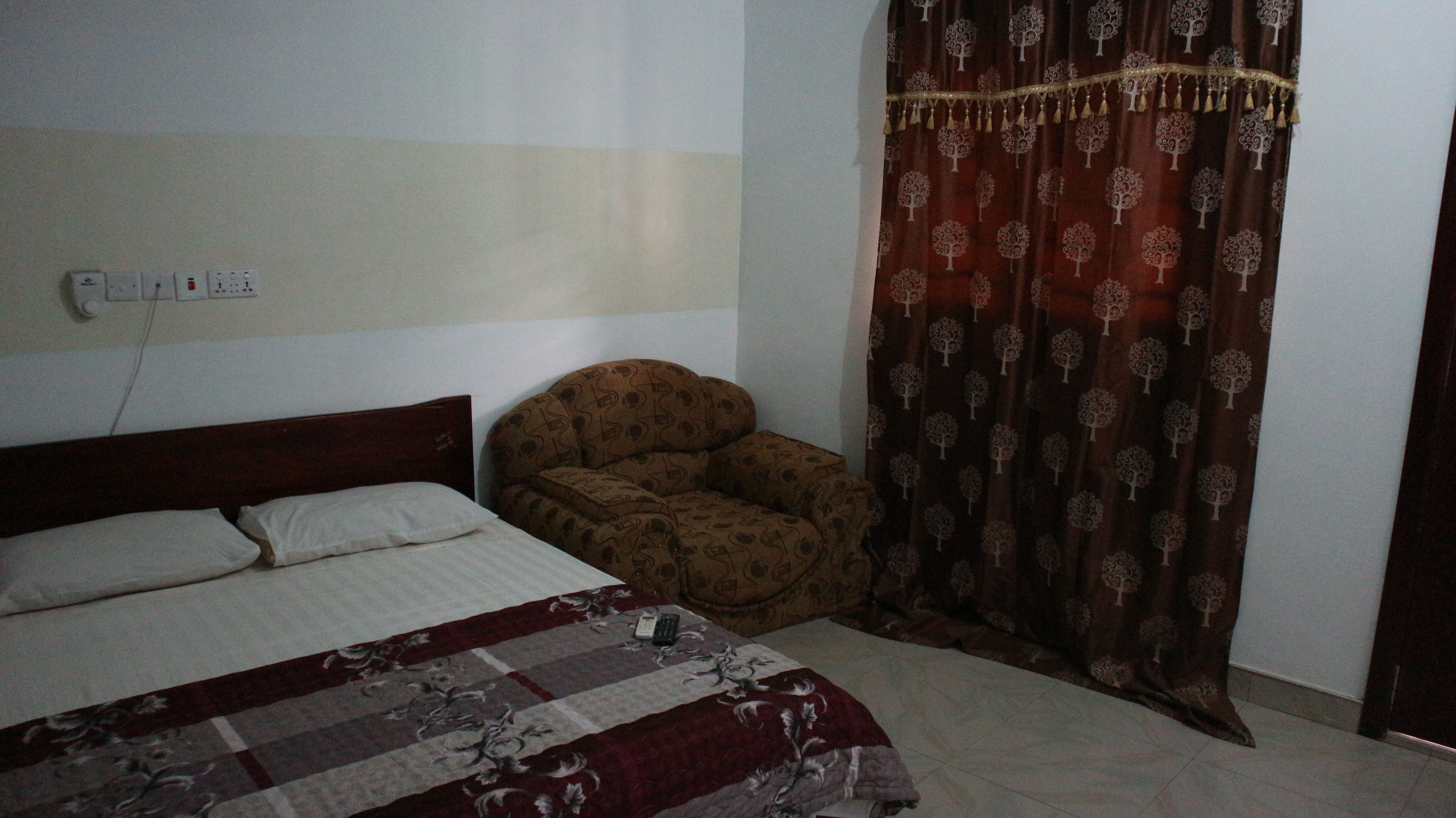A bedroom with a bed, a chair, a patterned curtain, and a wall socket.