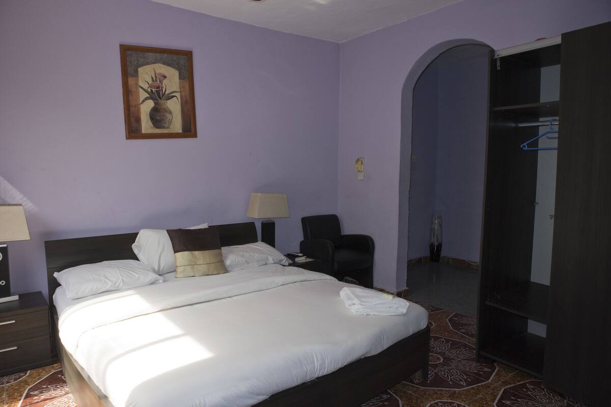 Habitació d'hotel amb un llit, dues taules de nit, una cadira, un armari i un quadre a la paret.