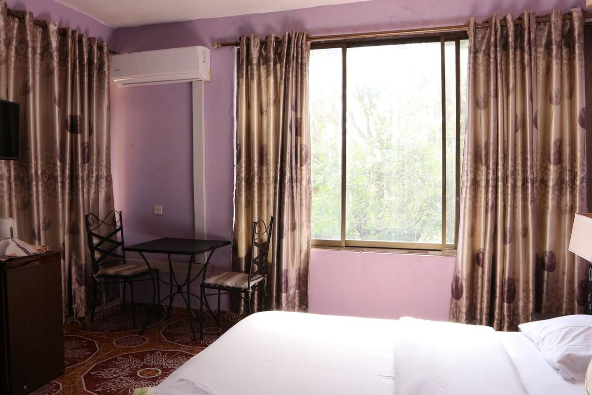 Una habitació d'hotel amb un llit, una taula, una cadira, aire condicionat i una finestra amb cortines.