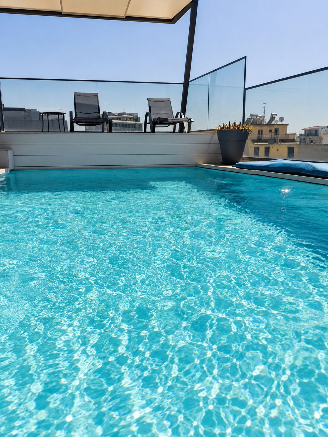 Une piscine sur le toit, avec une eau d’un bleu limpide, deux chaises longues, une plante en pot et une vue sur le paysage urbain.
