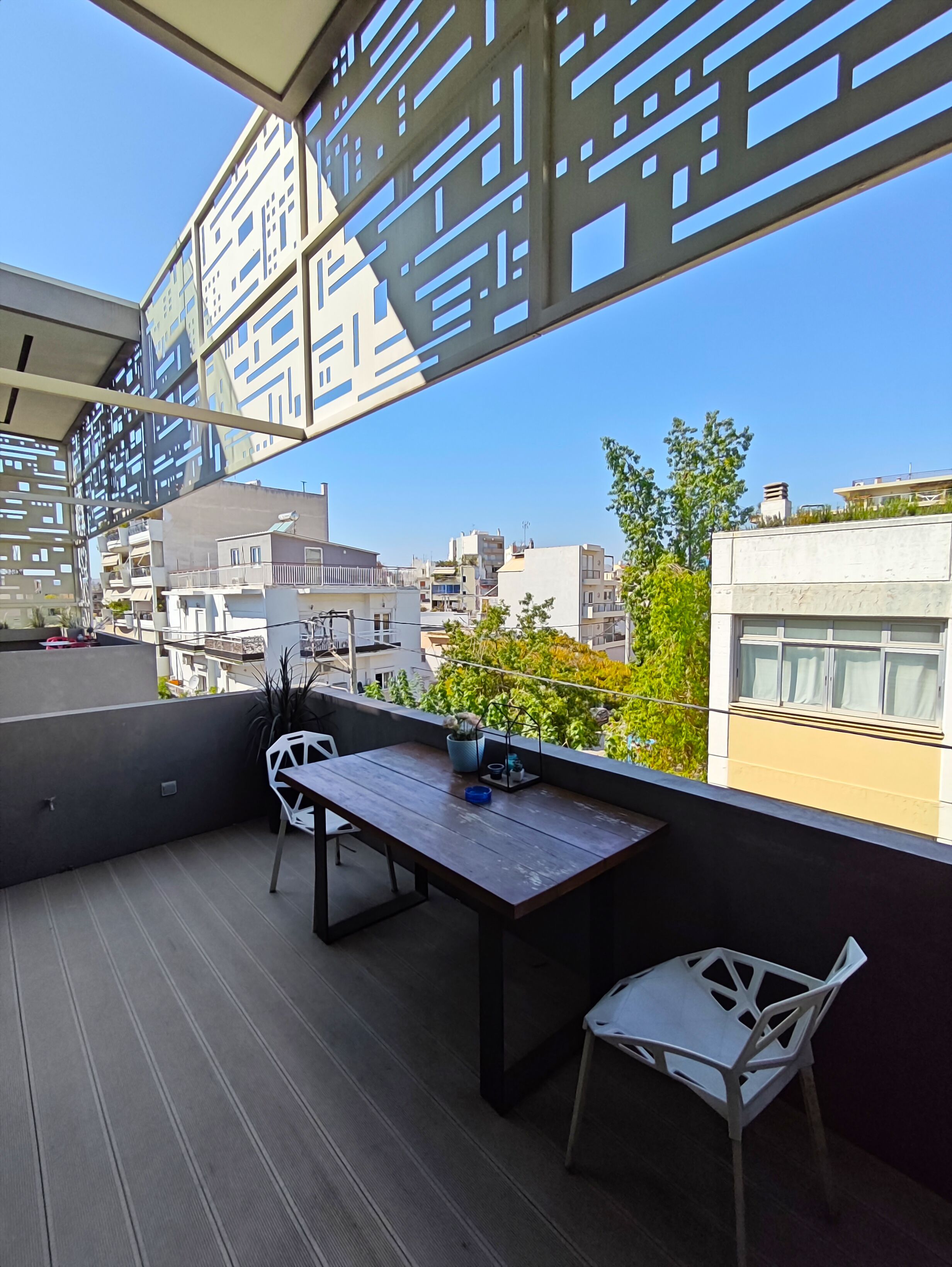 A balcony with a wooden table and chairs, a potted plant, and a view of surrounding buildings.