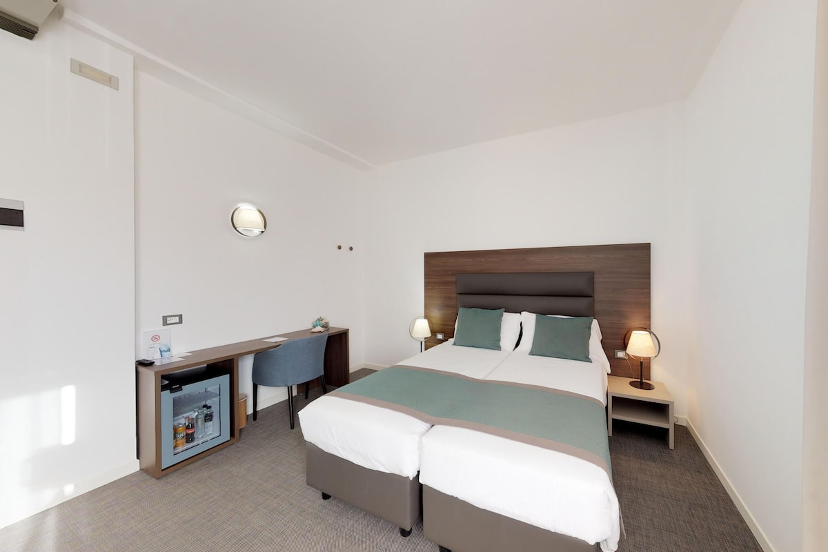 A hotel room with a bed, bedside tables, a desk, and a minibar.