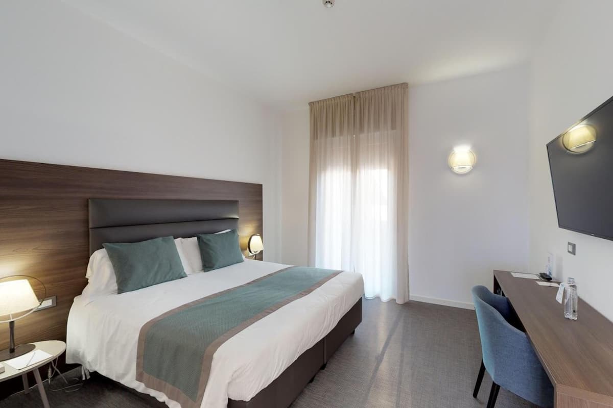 A hotel room with a bed, bedside lamps, a desk with a chair, a television, and a large window with curtains.