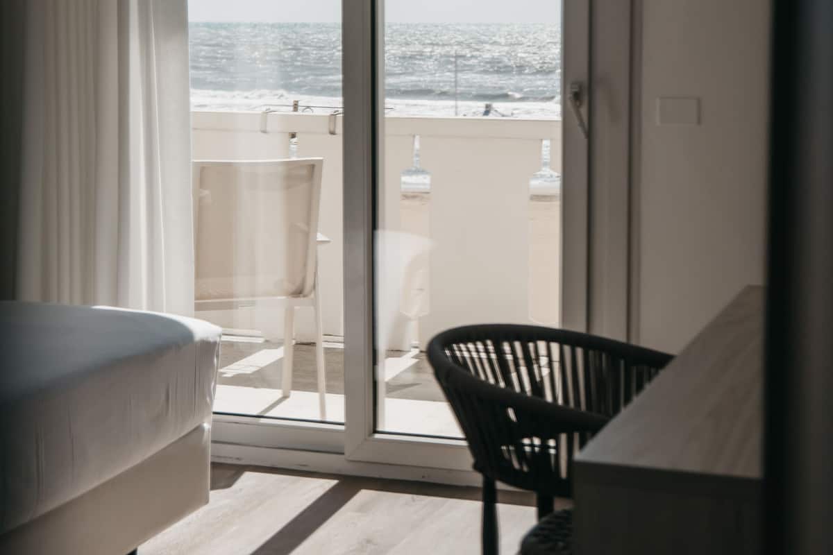 A room with a bed, a chair, and a sliding glass door leading to a balcony with a view of the sea.