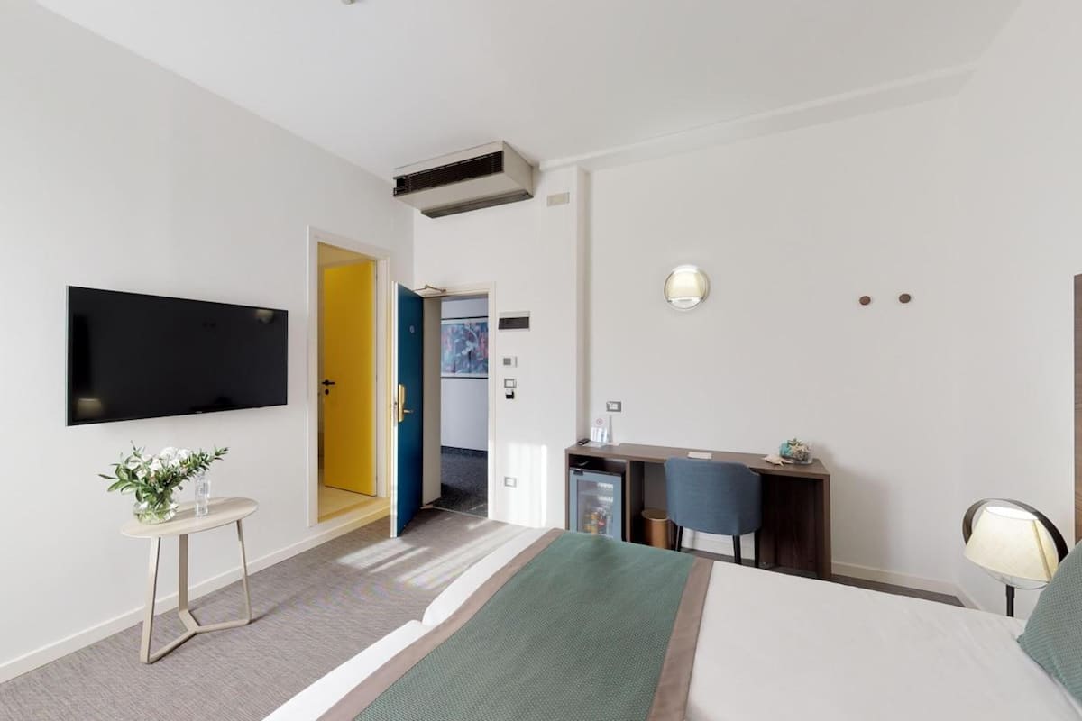 A hotel room with a bed, a desk, a television, a small table with a plant, and a door leading to another room.