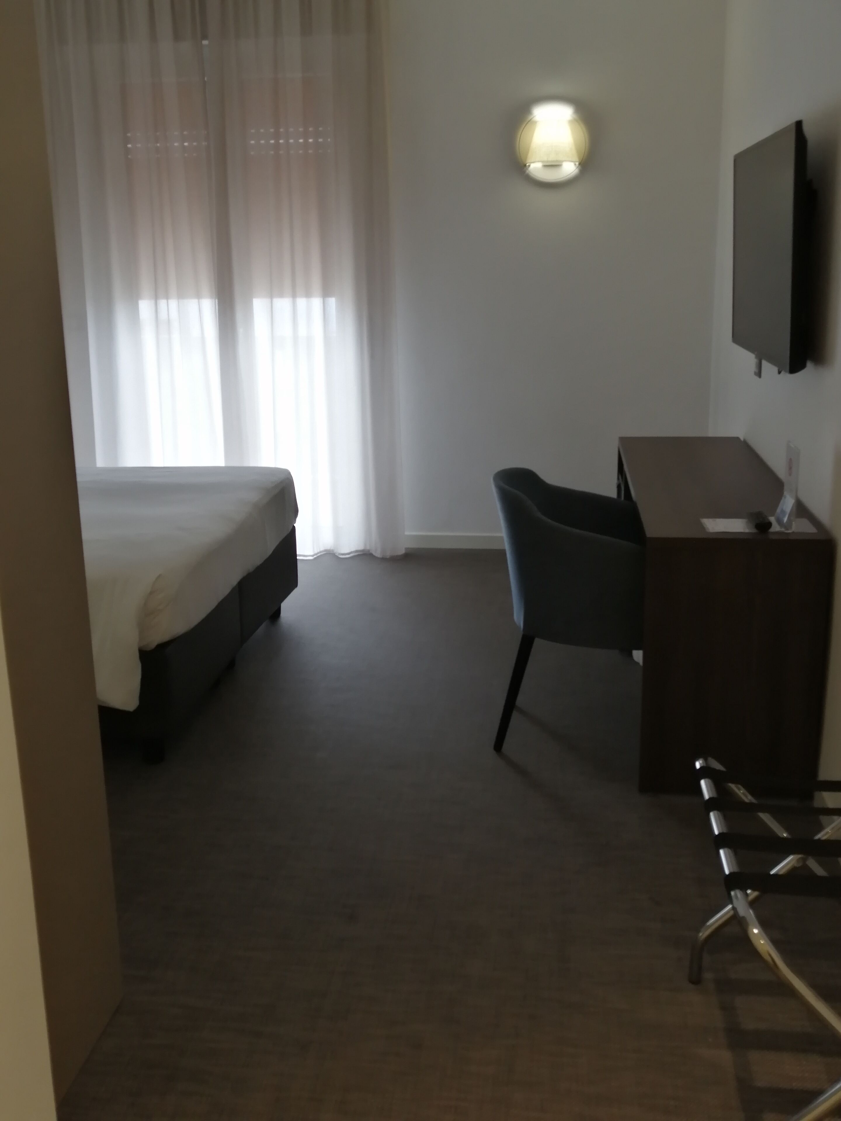 A hotel room with a bed, a desk, a chair, a flat-screen TV mounted on the wall, and a window with sheer curtains.