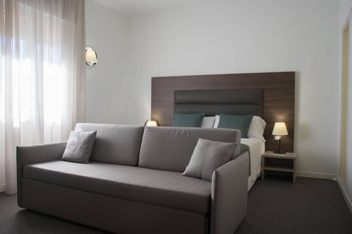 A hotel room with a grey sofa, a bed with a wooden headboard, two bedside tables with lamps, and sheer curtains on the window.