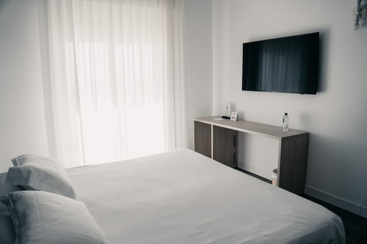 A hotel room with a large bed, a wooden desk, a flat-screen TV, and a window with sheer curtains.