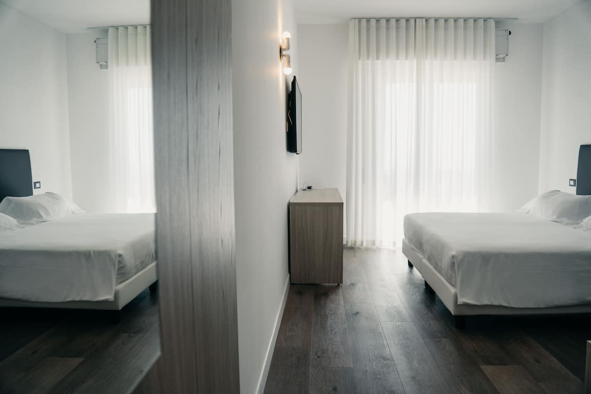A hotel room with two beds, a small wooden nightstand, a wall-mounted TV, and a window with curtains.