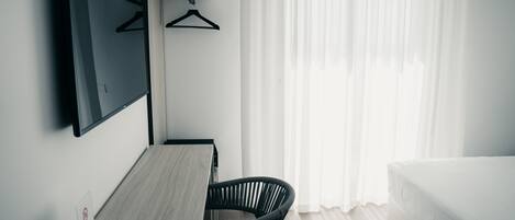 A hotel room with a bed, a desk with a chair, a TV mounted on the wall, and a window with sheer curtains.