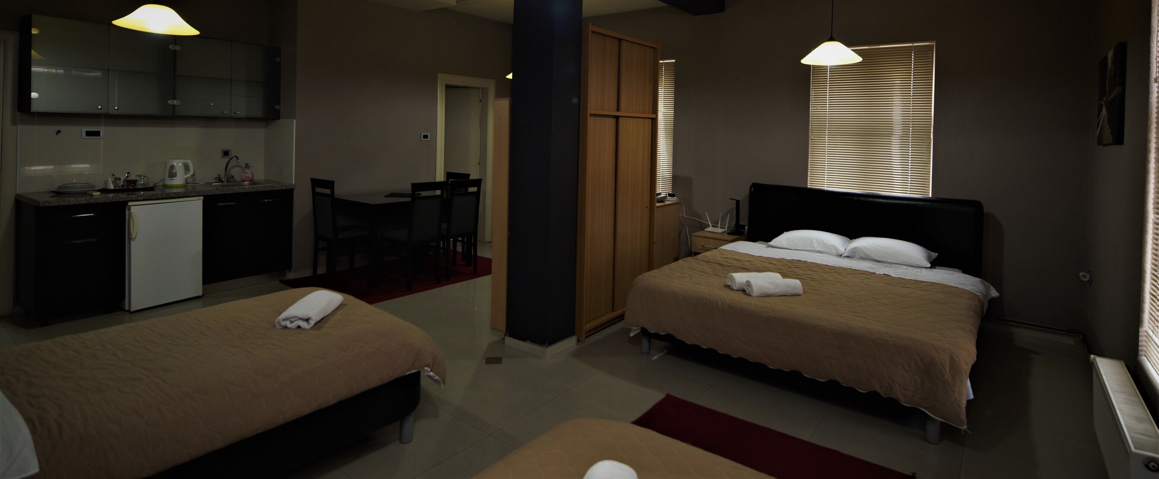 A hotel room with two beds, a kitchenette, a dining area, and a window with blinds.