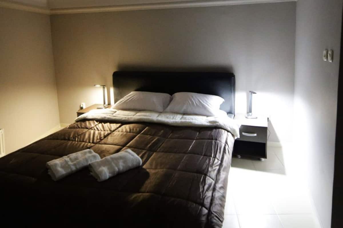 Una cama bien hecha con un edredón marrón oscuro, dos almohadas y una toalla doblada. Hay una mesita de noche con una lámpara y un pequeño dispositivo electrónico.