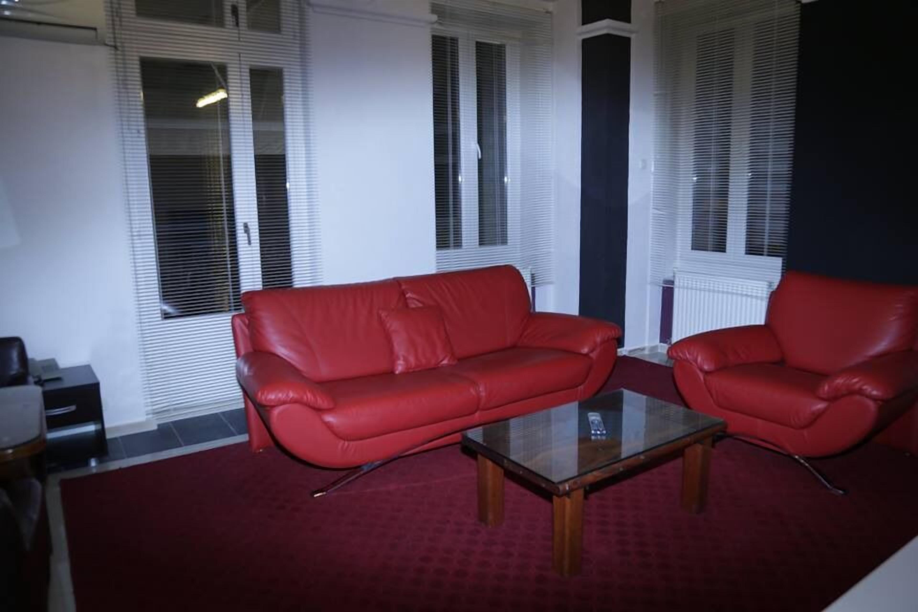 Un salon avec un canapé et un fauteuil en cuir rouge, une table basse en verre et des stores blancs aux fenêtres.