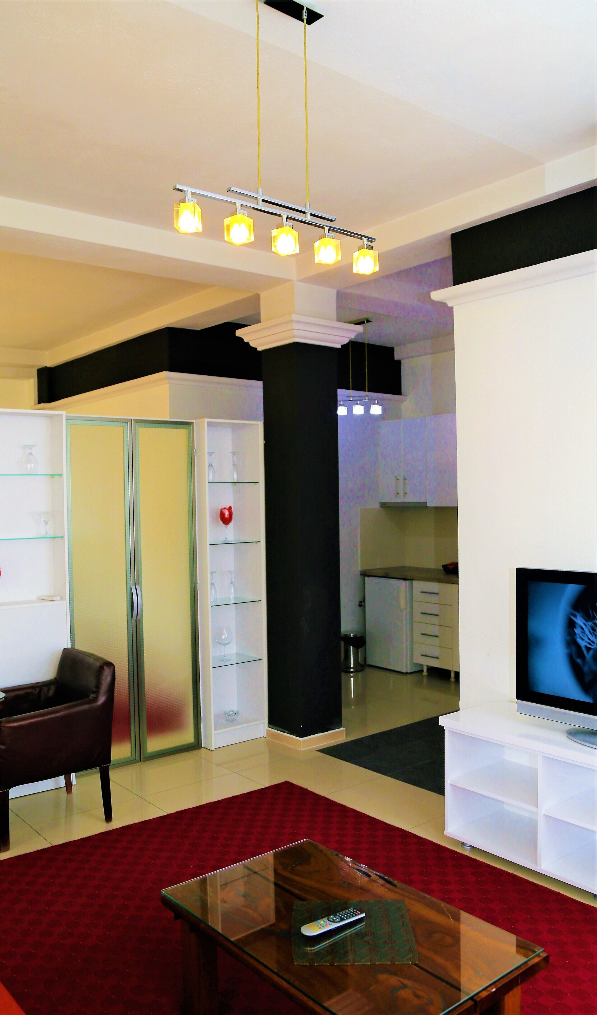 A modern living room with a glass coffee table, a red carpet, a television, and a kitchen area in the background.