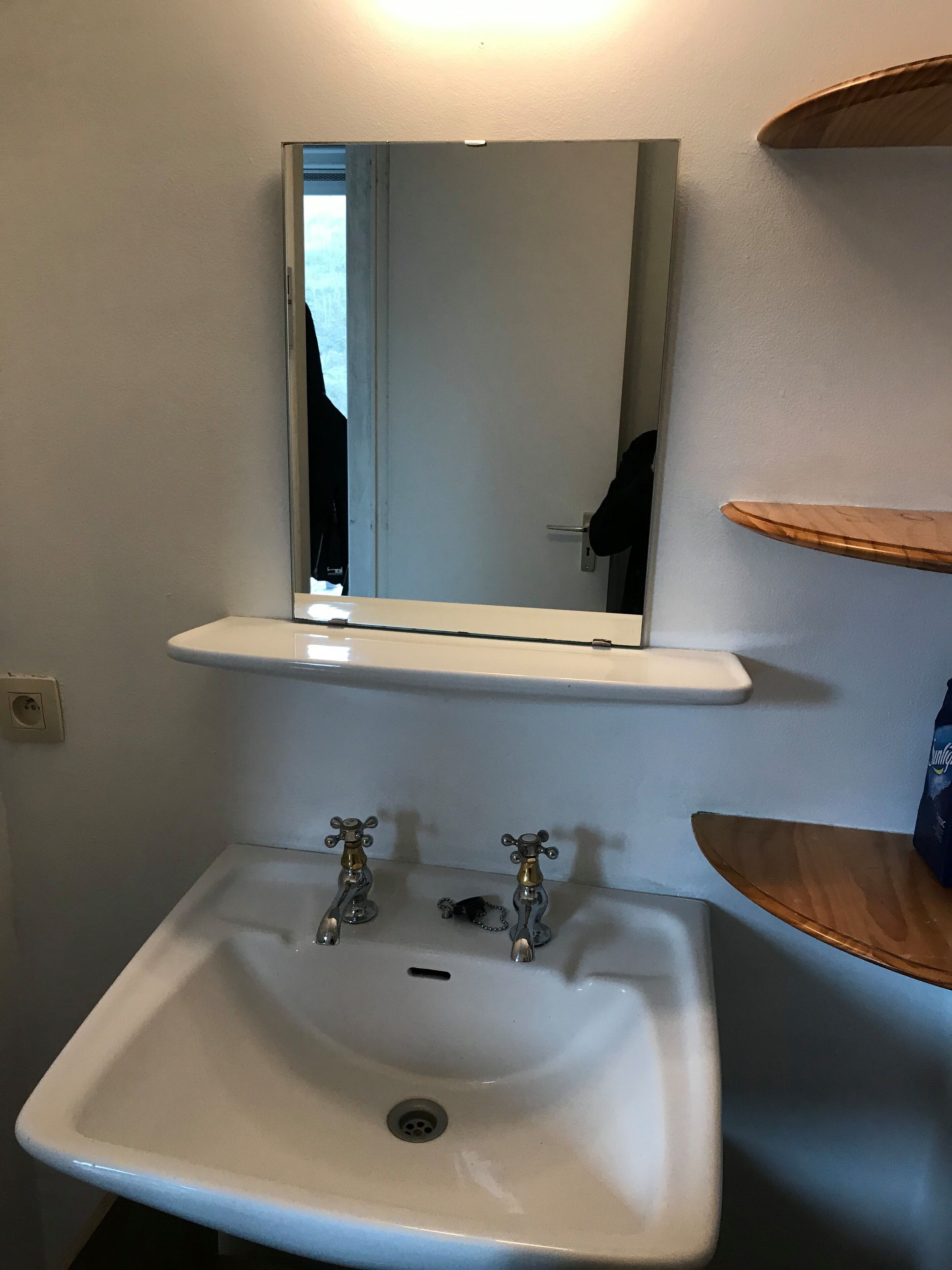 A bathroom with a white sink, a mirror above it, and a shelf with a bag of tissues.