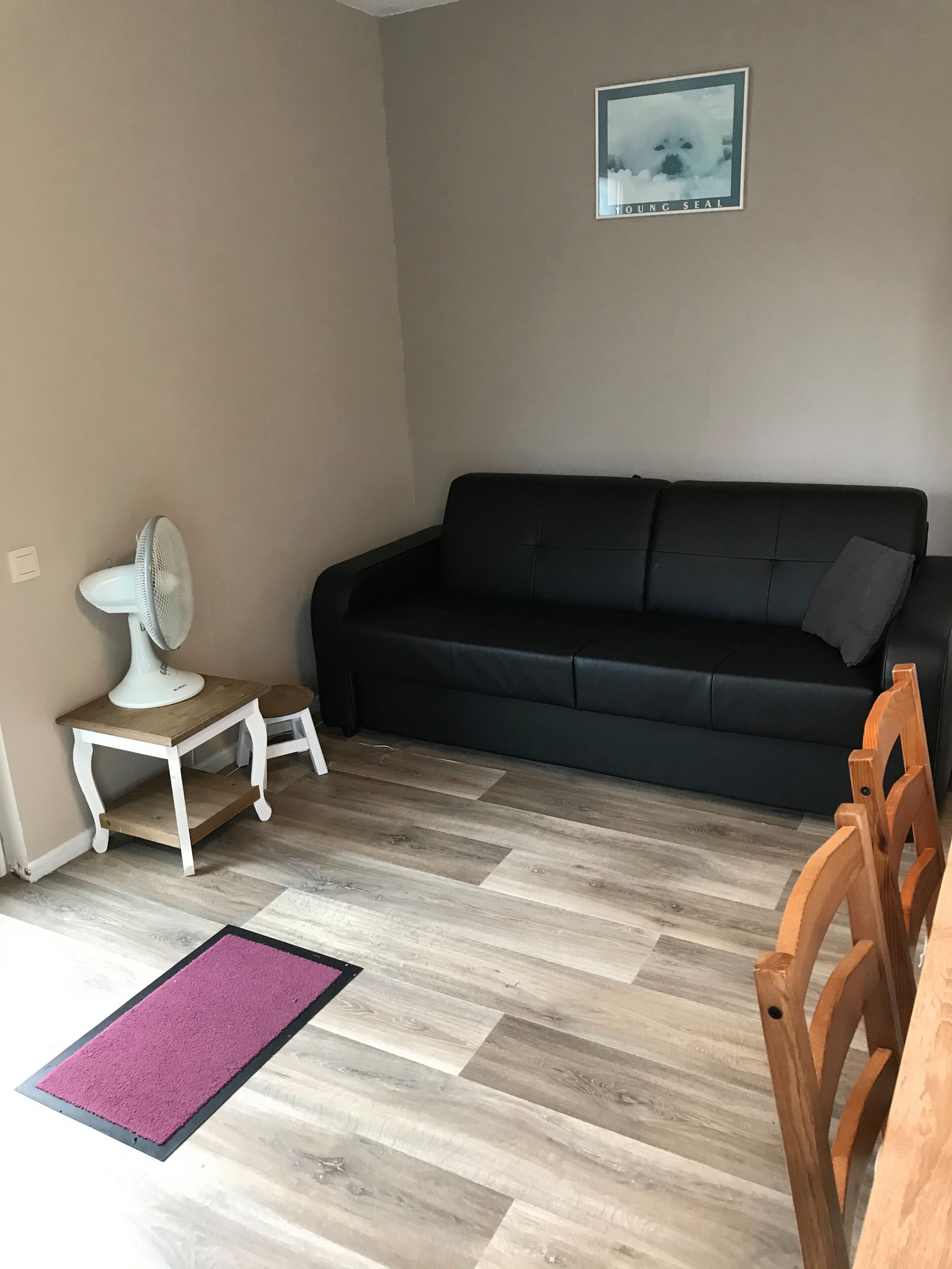A living room with a black sofa, a small white table with a fan, a purple rug, and a wooden chair.
