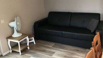 A living room with a black sofa, a small white table with a fan, a purple rug, and a wooden chair.