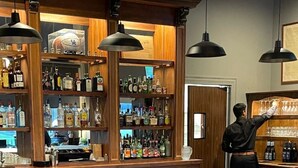 A bar area with wooden shelves stocked with bottles, a bartender preparing drinks, and patrons seated at tables. - The Meadows Hotel (Fulton)