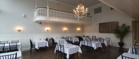 A dining area with multiple tables and chairs, a chandelier, and a balcony with railings.