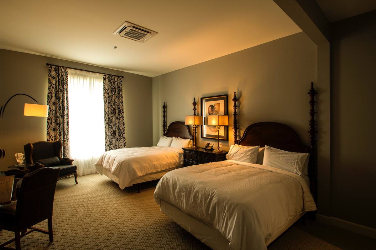A hotel room with two beds, a desk, a chair, and a lamp.