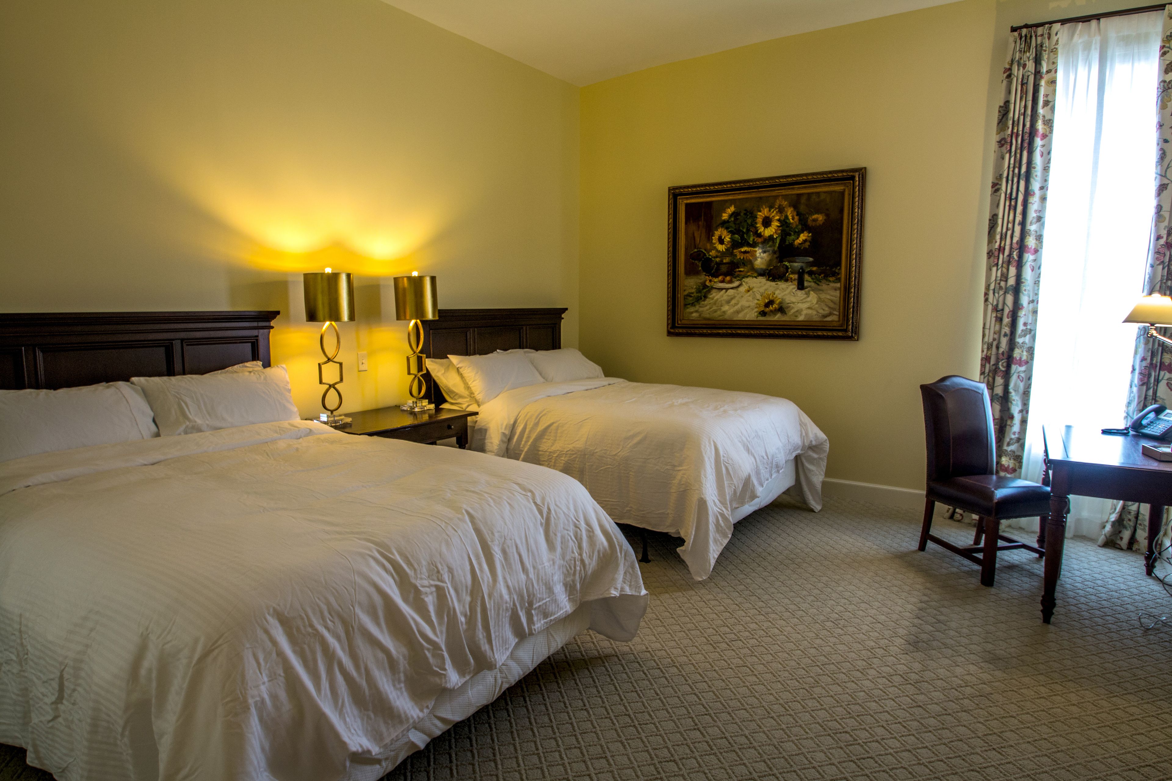 A hotel room with two beds, a desk with a chair, a painting on the wall, and a lamp on the nightstand.