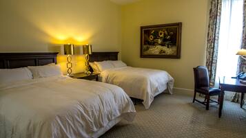 A hotel room with two beds, a desk with a chair, a painting on the wall, and a lamp on the nightstand.